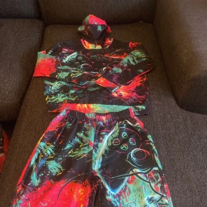 Färgstark hoodie med gamingtryck - Säljer en riktigt cool hoodie med gamingtema och färgstarkt mönster i svart, röd, grön och blå. Tröjan har huva och långärmad passform, med ett stort tryck av en spelkontroll på framsidan. Perfekt för dig som älskar gaming och vill sticka ut med din stil.