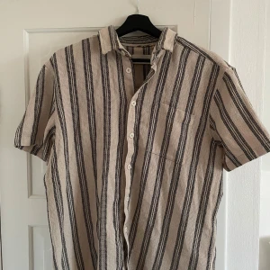 Randig kortärmad skjorta i bomull - Beige och svart randig skjorta i luftig bomull med korta ärmar och klassisk krage. Skjortan har knäppning framtill och en bröstficka. Perfekt för en avslappnad och stilren look.