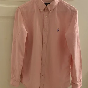 Rosa skjorta från Ralph Lauren XL - Snygg rosa skjorta från Ralph Lauren i storlek XL i barnstorlek men passar även S och M i herrstorlek, med klassisk button-down krage och broderad logga på bröstet. Skjortan har långa ärmar, custom fit och är gjord i mjuk bomull. Perfekt för dig som gillar stilrena och tidlösa plagg.