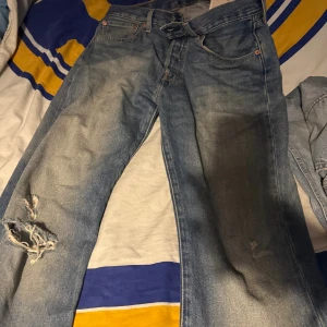 Levis 501 ljusblå jeans med slitningar - Ljusblå Levis 501 jeans med klassisk femficksdesign och slitna detaljer på vänster ben. Jeansen har rak passform och är tillverkade i robust denim. Perfekt för dig som gillar en avslappnad och vintage look.