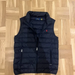 Mörkblå dunväst från Polo Ralph Lauren - Snygg mörkblå dunväst från Polo Ralph Lauren med klassisk quiltad design och röd logga på bröstet. Västen har två dragkedjefickor framtill och hög krage. Perfekt för lager-på-lager och stilren look.