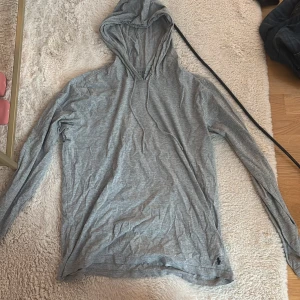 Grå hoodie från Polo Ralph Lauren - Snygg grå hoodie från Polo Ralph Lauren med klassisk huva och snörning. Tröjan har lång ärm och är tillverkad i mjuk bomull. Diskret Polo-logga broderad i mörkblått vid nederkanten. Perfekt för chill dagar och enkel att matcha.