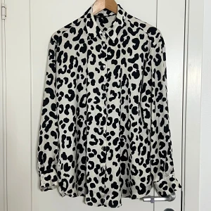 Leopardmönstrad skjorta från Zara - Snygg skjorta från Zara i storlek L med svart leopardmönster på vit botten. Skjortan har klassisk krage, knappar framtill och långa ärmar med manschett. Materialet är mjukt och har en lätt glansig finish som ger en cool vibe.