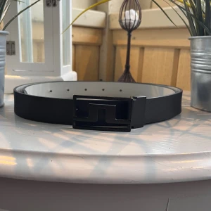 Svart skärp med stilren spänne - Snyggt svart bälte med en modern, rektangulär spänne i svart metall. Bältet har en slät yta och insidan är vit, vilket ger en cool kontrast. Perfekt accessoar för att lyfta din outfit med en minimalistisk touch.