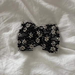 Svart och silver glittrigt hårspänne - Unikt hårspänne i svart och silver med glittriga pärlor som bildar ett snyggt mönster. Spännet har en böjd form och är dekorerat med små facetterade pärlor som ger extra bling. Perfekt för att ge frisyren en cool och trendig touch.