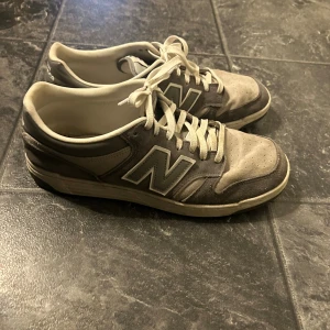 Grå New Balance sneakers mocka - Säljer ett par klassiska New Balance sneakers i grått med stora N-loggor på sidorna. Skorna har ovandel i mocka och textil, vita snören och en platt sula. Perfekta för dig som gillar en stilren och sportig look.