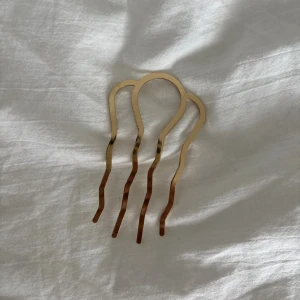 Guldfärgad hårkam med vågig design - Snygg hårkam i guldfärgad metall med tre vågiga pinnar. Perfekt för att skapa en cool och unik frisyr. Den minimalistiska och moderna formen gör den till ett statement i håret. Passar dig som gillar att sticka ut med detaljer.