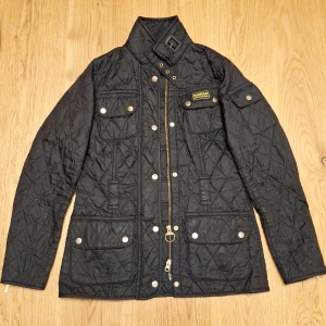 Svart quiltad jacka Barbour strl 34 - Quiltad svart jacka från Barbour International med cool ståkrage och spänne, flera fickor med tryckknappar och snygg gulddragkedja. Jackan har Barbour-märke på bröstet och är perfekt för dig som gillar klassisk stil med edge.