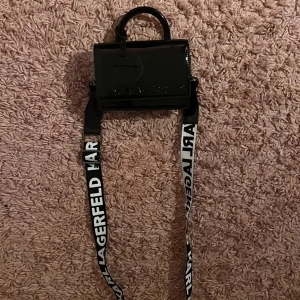 Svart handväska Karl Lagerfeld Kids - Cool svart handväska från Karl Lagerfeld Kids i glansigt syntetmaterial. Väskan har ett handtag upptill, avtagbar axelrem med logga i svartvitt och en silverfärgad Karl Lagerfeld-tag framtill. Perfekt för dig som vill sticka ut med en trendig accessoar.