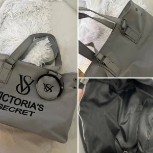 Grå shoppingväska Victoria's Secret - Stor grå från Victoria's Secret med svart logga framtill och matchande rund liten väska fäst vid handtaget. Väskan har breda handtag, silverfärgade detaljer och rymligt svart foder. Perfekt för dig som gillar stilren och praktisk design.