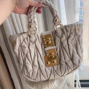 Beige handväska från Miu Miu i skinn - Authentic. Snygg beige handväska från Miu Miu med flätade handtag och coola guldfärgade metalldetaljer framtill. Väskan är quiltad och har ett rymligt fack med innerficka och dragkedja. Perfekt för dig som gillar lyxiga och unika accessoarer.