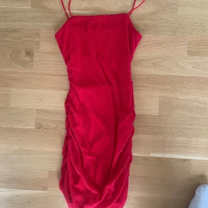 Röd glittrig kortklänning med smala axelband - Säljer en röd glittrig kortklänning med smala axelband och snygga rynkade detaljer längs sidorna. Klänningen har en figurnära passform och är tillverkad i ett stretchigt syntetmaterial som skimrar i ljuset. Perfekt för dig som vill sticka ut med en färgstark look.