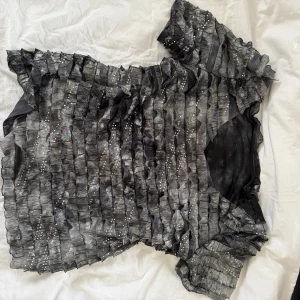 Glittrig svart blus med volangdetaljer - Säljer en unik svart blus med glittriga paljettdetaljer och volangiga lager över hela toppen. Blusen har korta ärmar och en lätt transparent känsla. Passar från S-L