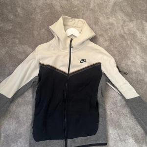 Nike tech tröja  - Nike hoodie med dragkedja i vit, svart och grå. Jackan har hög krage, svart Nike-logga på bröstet och en snygg detalj med dragkedjeficka på ärmen. 