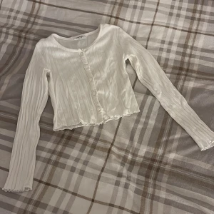 Vit ribbad blus med knappar - Säljer en söt vit ribbad blus från Kappahl med långa ärmar och knappar framtill. Toppen är croppad med vågig kant nertill och har en rund halsringning. Perfekt att styla med jeans eller kjol för en trendig look.