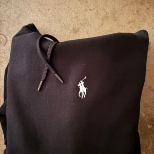 Svart hoodie från Polo Ralph Lauren - Svart hoodie från Polo Ralph Lauren med klassisk broderad logga på bröstet. Tröjan har huva med snörning och en stor magficka. Mjuk och skön bomullskvalitet, perfekt för chill dagar. Snygg och enkel design som alltid funkar.