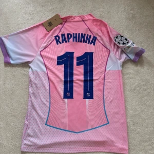 Barça Raphinha rosa matchtröja Nike Size S - Säljer en FC Barcelona fotbollströja med Raphinha och nummer 11 på ryggen. Tröjan är rosa med vita och blå detaljer, har korta ärmar och är tillverkad i lätt polyester. Snygga ränder, holografiskt Nike-märke och klubbemblem framtill. Champions League-patch på ärmen.