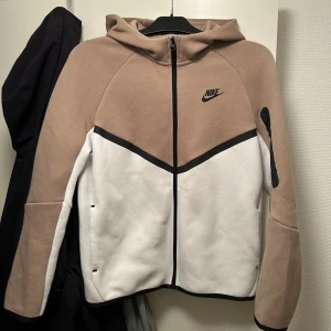 Nike Tech - Nike hoodie i beige och vitt med svart logga på bröstet. Tröjan har hel dragkedja, huva och svarta detaljer längs dragkedjan och ärmen. Tillverkad i mjuk bomullsblandning, perfekt för chill eller träning. Snygg och sportig look med lång ärm och praktiska fickor.