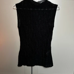 Svart spetsig ärmlös topp från Zara - Svart ärmlös topp från Zara i storlek S, tillverkad i ett tunt och genomskinligt spetsmaterial. Toppen har en rak passform och hög hals, med snyggt mönstrad spets över hela plagget. Perfekt för att skapa en cool och elegant look. Har endast testat den 🌟