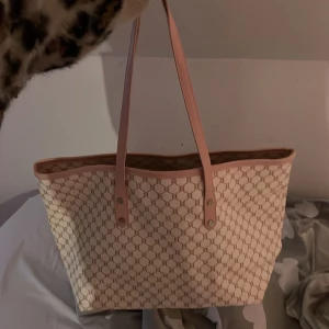 Beige shoppingväska med rosa detaljer - Stor shoppingväska i beige med ljusrosa handtag och kanter. Väskan har ett diskret mönster i guldton och stängs med dragkedja. Invändigt finns ett ljust foder och en innerficka med dragkedja. Perfekt för dig som vill ha plats för allt du behöver under dagen.