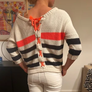 Randig stickad tröja med snörning - Säljer en cool stickad tröja med breda marinblå och beige ränder samt en stark orange rand framtill. Baksidan har snygg snörning med orange band och tröjan har trekvartsärm. Passformen är loose och materialet känns mjukt och grovstickat.