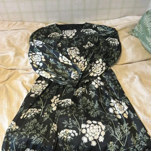 Blommig långklänning från Stockh LM - Snygg långklänning med svart botten och blommigt mönster i vitt och grönt från Stockh LM. Klänningen har långa ärmar, knappar framtill och ett mjukt, flowigt tyg som ger en chill vibe. Använd en gång på en begravning, storlek S men passar som M. Nypris 500kr 