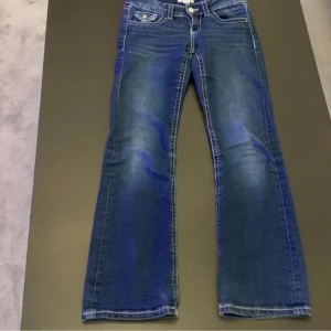 Blå bootcut jeans med ficklock - Snygga blå jeans med bootcut passform och coola ficklock med knapp både fram och bak. Jeansen har kontrastsömmar och klassisk femficksdesign. Materialet är jeans med en mörkblå tvätt och lätt slitning på låren. Perfekt för dig som gillar retrovibbar och detaljer.