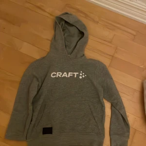 Grå hoodie från Craft med logga - Säljer en grå hoodie från Craft med vit logga framtill. Tröjan har huva, känguruficka och är tillverkad i mjukt material som passar perfekt för chill eller träning. Enkel och sportig stil som funkar året runt.