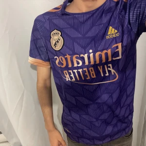 Real Madrid Ronaldo 7 lila matchtröja - Säljer en snygg Real Madrid fotbollströja från Adidas i lila med guldiga detaljer och tryck. Tröjan har korta ärmar, mönstrad design och Ronaldo 7 på ryggen. Materialet är lätt och andas, perfekt för träning eller match. Officiell klubbmärke på bröstet.