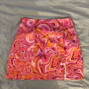 mönstrad kortkjol från New Yorker - Säljer en färgstark kortkjol från New Yorker i storlek XS med psykedeliskt mönster i rosa, orange och rött. Kjolen har en liten slits framtill och är gjord i lätt syntetmaterial