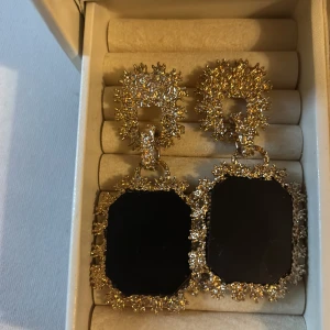 Statement örhängen med svart och guld - Stora statement örhängen med rektangulär svart sten och glittrig, detaljerad guldram. Örhängena har en unik, nästan spikig kant och ger en lyxig känsla. Perfekta för dig som vill sticka ut och addera extra bling till din look.
