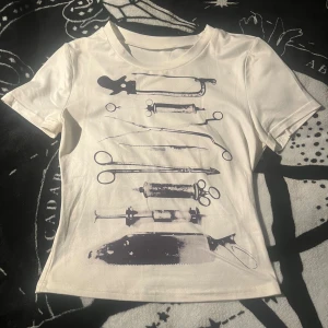 Vit t-shirt med kirurgiska verktyg - Vit kortärmad t-shirt med tryck av svarta kirurgiska verktyg på framsidan. Tight passform och rund halsringning. Perfekt för dig som gillar edgy och unika motiv.