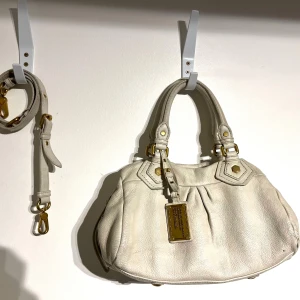 Vit handväska från Marc by Marc Jacobs - Snygg vit handväska i äkta skinn från Marc by Marc Jacobs med guldfärgade metalldetaljer och logoplatta. Väskan har två handtag, avtagbar axelrem och dragkedja. Insidan är fodrad med svartvitt mönster med märkets logga. Finns tecken på användning och en skråma förekommer (se bild). Rekommenderar att använda en magic sponge på väskan eftersom att det texturerade lädret gör det enkelt för smuts att sätta sig i springor. Mått: 30x22x12cm, Axelrem medföljer.  