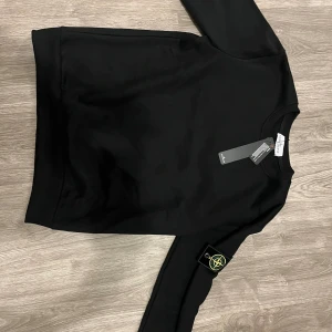 Svart sweatshirt från Stone Island - Svart sweatshirt från Stone Island med klassisk rund hals och ribbade muddar. Tröjan har den ikoniska Stone Island-loggan på vänster ärm och är tillverkad i mjuk bomull. Perfekt för dig som gillar stilrena och trendiga plagg.