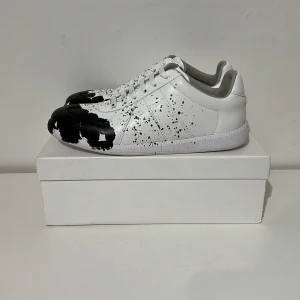 Maison Margiela svartvit sneakers - Maison Margiela sneakers i vitt skinn med svart färgstänk-design över tån och sidorna. Klassisk låg modell med snörning och rund tå. Snygg kontrast mellan det vita och svarta, och den unika splatter-looken gör dem riktigt feta. Kommer med originalkartong.