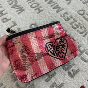 Paljettkuvertväska från Victoria's Secret - Snygg kuvertväska med paljetter i rosa och röda toner, rutigt mönster och svart dragkedja. Framsidan har ett hjärta med texten 'LOVE VICTORY'. Baksidan är svart med rosa Victoria's Secret-logga. Perfekt för att addera lite extra glitter till din look.