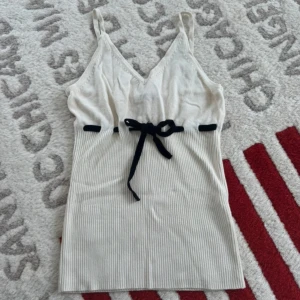 Vit ribbad linnetopp från Gina Tricot - Säljer en vit ribbad linnetopp från Gina Tricot med smala axelband och v-ringning. Toppen har en svart knytning i midjan som ger en snygg detalj. Perfekt för varma dagar och enkel att styla med jeans eller kjol.