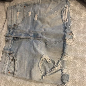 Levi's ljusblå jeansshorts med fransar - Säljer ett par ljusblå jeansshorts från Levi's med råa, fransiga kanter och slitna detaljer både fram och bak. Klassisk femficksmodell med knappgylf 