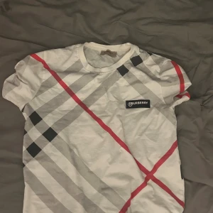 Vit rutig Burberry t-shirt - Snygg vit t-shirt från Burberry med klassiskt rutigt mönster i svart, grått och rött. T-shirten har korta ärmar och rund halsringning. Ikonisk Burberry-logga på bröstet. Perfekt för dig som gillar stilren design med en twist.