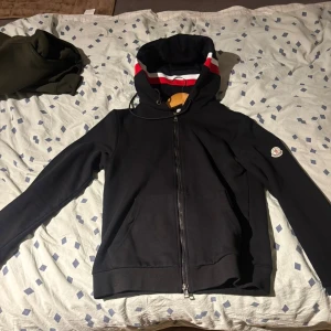 Svart Moncler hoodie - Svart hoodie från Moncler med hel dragkedja och huva. Insidan av huvan har snygga ränder i rött, vitt och svart. Tröjan har Moncler-logga på ärmen och praktiska fickor framtill. Perfekt för dig som gillar stilrena och exklusiva streetwear-plagg.