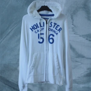 Vit hoodie från Hollister med blå tryck - Vit hoodie från Hollister med blå text och siffror på bröstet. Tröjan har huva med snörning, dragkedja framtill och två fickor. Långärmad och tillverkad i mjuk bomullsmix, perfekt för chill dagar. Klassisk college-vibe och snygg passform.