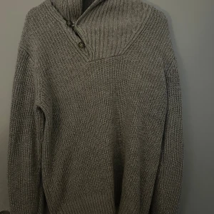 Grå stickad polotröja från L.O.G.G. - Säljer en grå stickad polotröja från L.O.G.G. by H&M med ribbad krage och två dekorativa knappar vid halsen. Tröjan har långärmad design och grovstickad struktur som ger en chill och varm vibe. Perfekt för kalla dagar och snygg att styla med jeans.