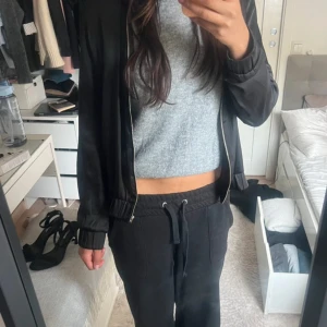 Svart bomberjacka i satin - Säljer en svart bomberjacka med stilren design och glansig satinfinish. Jackan har ribbade muddar vid ärmslut och nederkant, samt dragkedja framtill. Långärmad och med klassisk bomberkrage, perfekt för dig som gillar en clean och chill look.