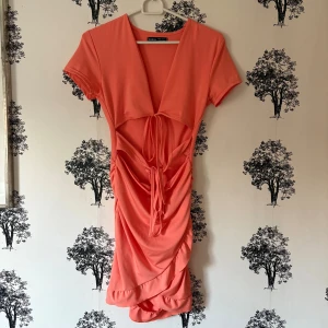 Korallrosa kort klänning från SHEIN - Superfin korallrosa kort klänning från SHEIN med djup v-ringning, cut-out detaljer och knyt framtill. Klänningen har korta ärmar, rynkade partier och volang nertill. Perfekt för dig som vill sticka ut med en färgstark look.