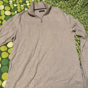 Beige stickad half zip från Jack & Jones - Säljer en beige stickad tröja från Jack & Jones med half zip och hög krage. Tröjan har långärmad design och är perfekt för dig som gillar enkel och stilren look. Passar dig som vill ha en chill och snygg vardagsstil.