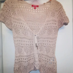 *INTRESSEKOLL* - Supersöt beige rosa stickad blus från Guess med korta ärmar och v-ringning. Blusen har ett snyggt hålmönster och knyt framtill, samt en liten metallbricka med Guess-logga i nacken. Perfekt till jeans eller kjol för en chill look.