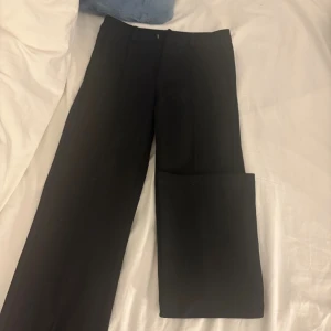 Svarta kostymbyxor från Kappahl - Snygga svarta kostymbyxor från Kappahl i rak modell. Byxorna har klassisk midja, bälteshällor och pressveck framtill för en clean look. Perfekta till en stilren outfit och passar dig som gillar enkel och tidlös design.