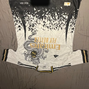 Real Madrid x Adidas draktröja S - Snygg Real Madrid fotbollströja från Adidas i vitt och svart med guldiga detaljer. Unikt drakmönster över hela tröjan, svart och guld klubbmärke, samt Emirates Fly Better-tryck. Tillverkad i lätt och glansigt material, perfekt för match eller träning.
