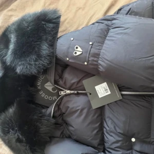 Svart Moonstone Parka från Moose Knuckles - Säljer en svart Moonstone Parka från Moose Knuckles med fluffig päls runt kragen och quiltad design. Jackan har dragkedja framtill, långa ärmar och coola metalldetaljer på ärmen. Perfekt för kalla vinterdagar och riktigt snygg streetstil.