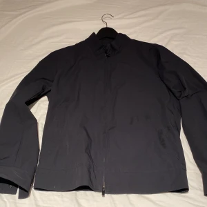 Svart vindjacka från Massimo Dutti S - Stilren svart vindjacka från Massimo Dutti i storlek S. Jackan har minimalistisk design, hög krage med tryckknappar och dragkedja framtill. Tillverkad i ett lätt och smidigt syntetmaterial, perfekt för blåsiga dagar. 